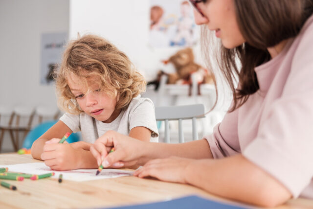 Close up of a child with an autism spectrum disorder and the therapist by a table drawing with crayons during a sensory integration session.
